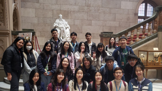 Group of young people posing indoors with a historic statue and ornate staircase in the background