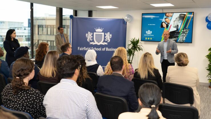 Speaker addressing audience at Fairfield School of Business event, room with digital screen and branded banner