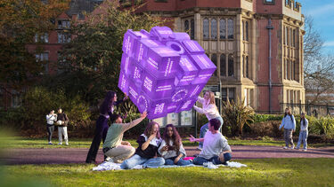 People sitting on grass interacting with a large purple cube outside a historic university building
