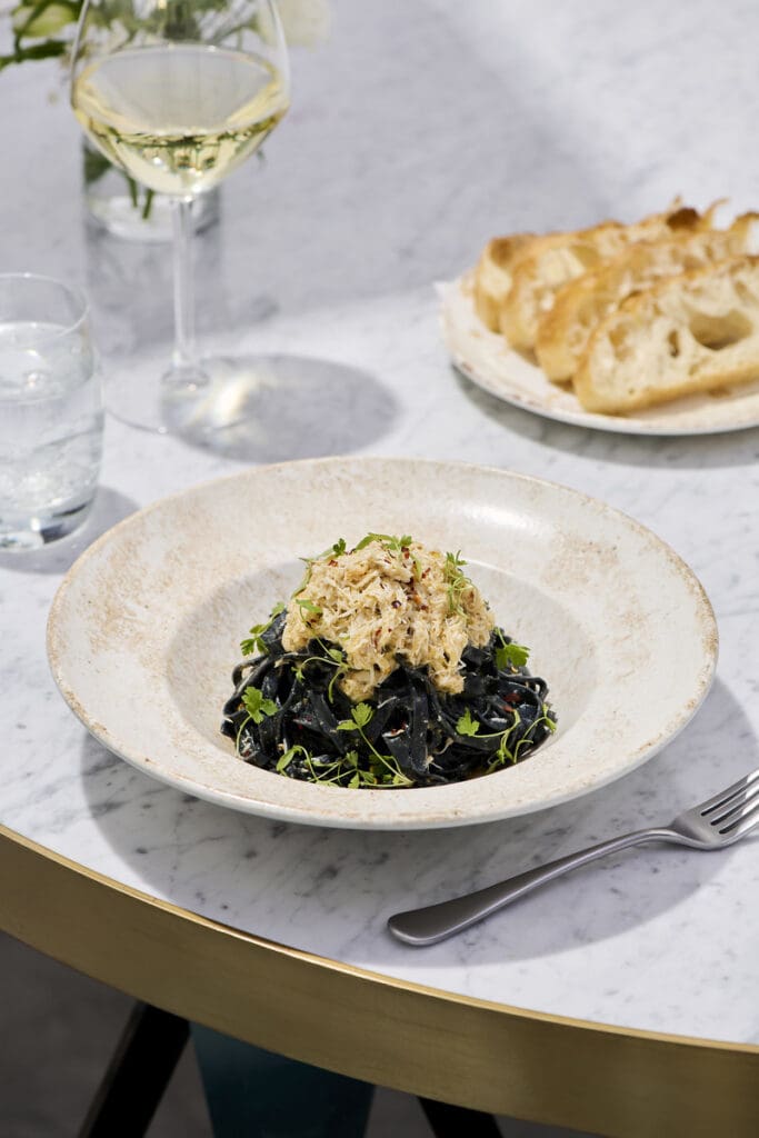 Black squid ink pasta with crab meat in a cream bowl, served with bread and a glass of white wine on a marble table