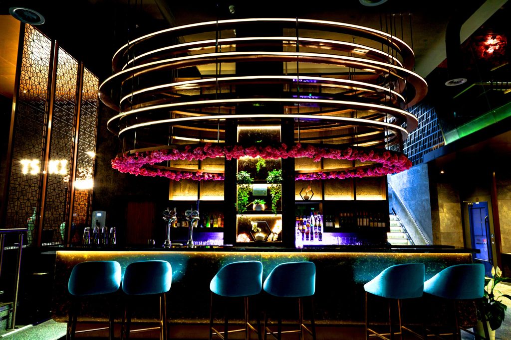 Modern bar interior with stylish lighting, blue chairs, floral decor, and a well-stocked shelf for an elegant atmosphere