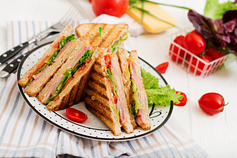 Plate of club sandwiches with ham, lettuce, and tomatoes on a striped cloth, surrounded by fresh ingredients