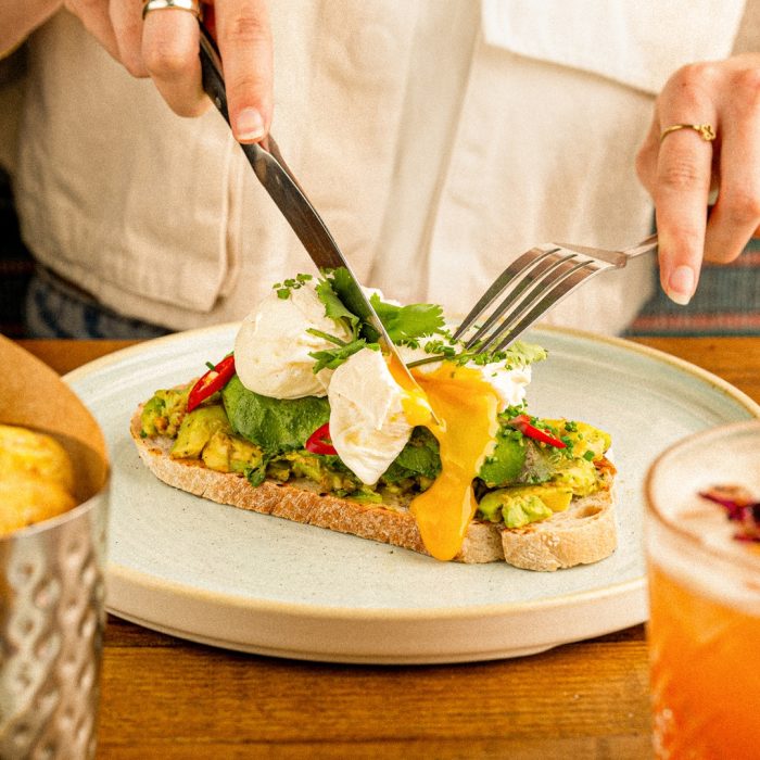 Person slicing avocado toast topped with poached eggs and herbs, next to a drink, in a cosy cafe setting