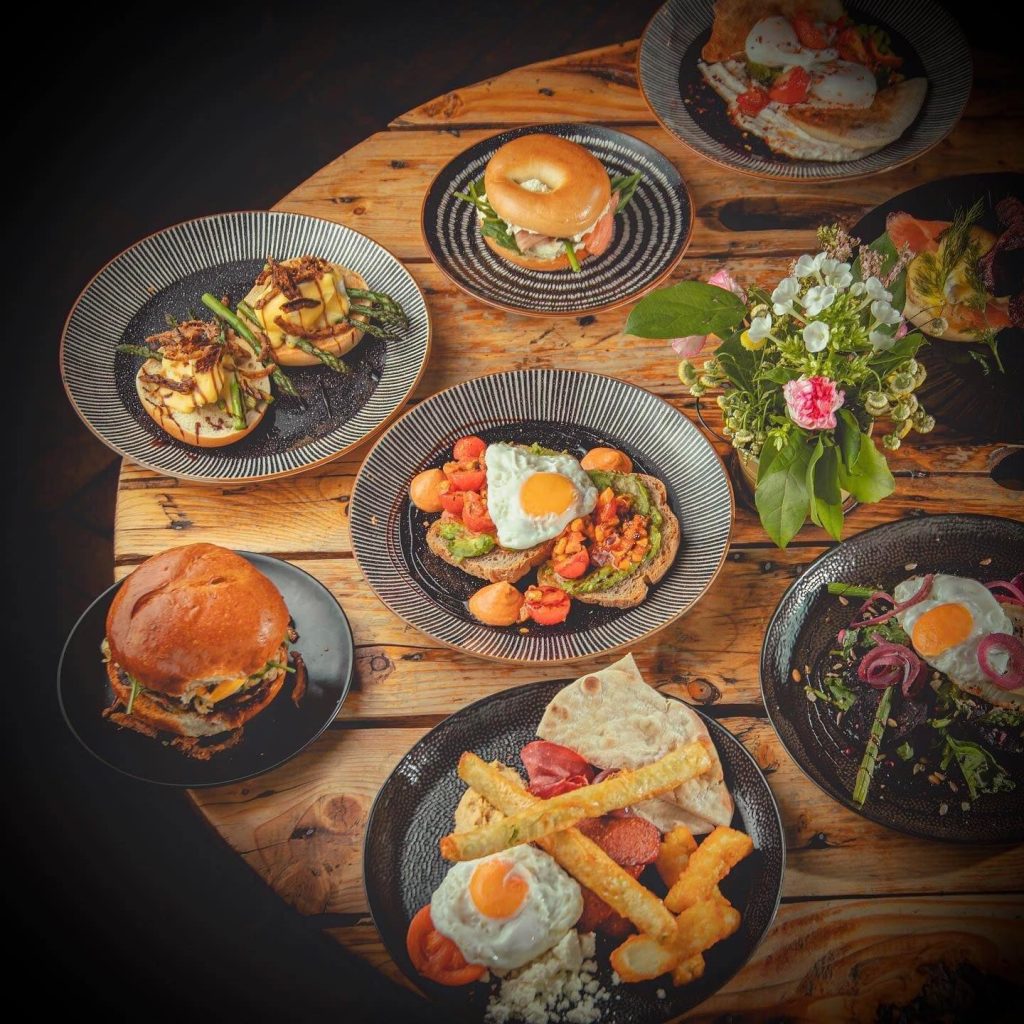 Assorted breakfast dishes on a rustic wooden table, featuring eggs, bagels, and vibrant salads with a floral centrepiece