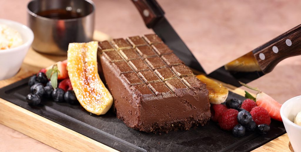 Decadent chocolate dessert with fresh berries and caramelised banana on a serving board being sliced by knife