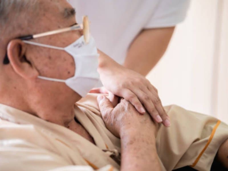 Elderly man wearing mask comforted by caregiver with supportive hand on shoulder, promoting care and compassion