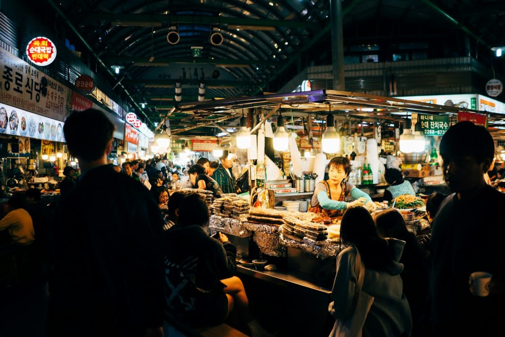 Bustling night market with diverse food stalls, vibrant lights, and people enjoying local delicacies under a covered roof